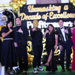 A group of nine people, dressed formally, pose in front of a gold and black balloon backdrop that reads "Unmasking a Decade of Excellence ProfitMasters.