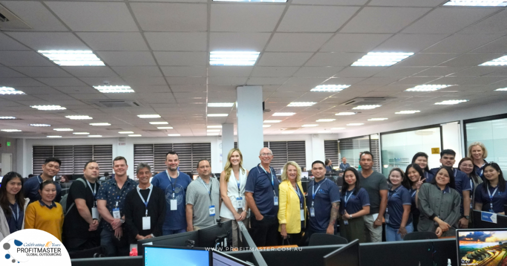 A large group of people, wearing company lanyards, pose for a photo in an office with desks and monitors. There is a banner in the bottom left corner with the text "ProfitMaster Group.