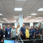 A large group of people, wearing company lanyards, pose for a photo in an office with desks and monitors. There is a banner in the bottom left corner with the text "ProfitMaster Group.