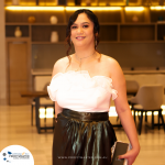 A woman in formal attire stands indoors, holding a phone. She is dressed in a white ruffled top and a dark skirt. The background features warm lighting and shelves with decorative items.