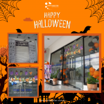 Halloween-themed office decorations with hanging witch and skeleton cutouts, orange and black balloons, and a "Happy Halloween" banner. "Wanted" poster decor and company branding are visible.