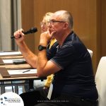 A man in a navy shirt is holding a microphone and resting his chin on his hand, sitting at a desk with a woman beside him. A "ProfitMaster" banner is visible in the bottom corner of the image.