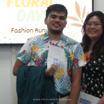 Two people standing in front of a "Floral Day" fashion runway sign, both smiling and holding papers.