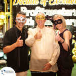 Three people wearing masks and formal clothing pose with thumbs up in front of a decorated backdrop celebrating a "Decade of Excellence" for Profitmaster's anniversary event.