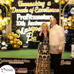 A man and a woman, both wearing gold masks, stand in front of a backdrop at a masquerade ball celebrating "Profitmaster's 10th Anniversary". The setting includes gold and black balloon decorations.