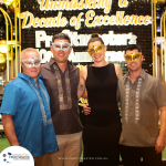 Four people in decorative masks are standing together, smiling in front of a backdrop with the text "Unmasking a Decade of Excellence" at an anniversary event.