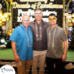 Three men wearing decorative masks and traditional barong tagalog shirts pose in front of a "Decade of Excellence" event backdrop adorned with balloons and celebratory decorations.