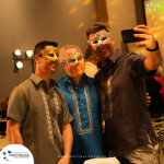 Three men wearing decorative masks take a selfie together at an indoor event.