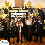 A group of seven people wearing formal attire and masks posing in front of a decorated backdrop that reads "Unmasking a Decade of Excellence, Profitmasters 10th Anniversary.