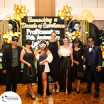 A group of seven people in semi-formal attire and masks stand together at an event with the backdrop reading "Unmasking a Decade of Excellence, Profitmaster’s 10th Anniversary Celebration.