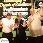 Three men wearing masquerade masks pose with hands outstretched at Profitmaster's 10th Anniversary celebration. A banner and balloons are visible in the background.