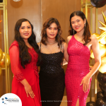 Three women in formal dresses pose together in front of a decorated backdrop with golden and black balloons and a sign that reads "PROFITMASTER.