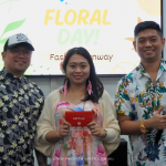 Three people in floral attire stand in front of a sign reading "Floral Day Fashion Runway," with one holding a red envelope.