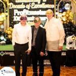 Three men wearing masks stand together in front of a decorated backdrop at an event with golden balloons. The backdrop reads "Celebrating a Decade of Excellence - Profitmaster 100 Million.