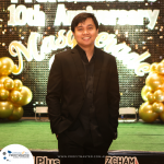 A person wearing a black suit stands smiling in front of a sparkly "10th Anniversary Masquerade" sign with gold balloons decorating the background.