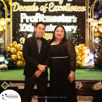 A man and a woman stand in formal attire in front of a decorated backdrop celebrating Profitmaster's 10th anniversary. The backdrop is adorned with gold and black balloons.