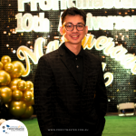 A person in a black suit stands in front of a decorated backdrop for the 10th anniversary of Profitmaster. The background features gold balloons and celebratory signage.