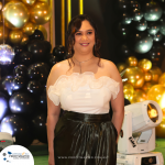 A woman in a white ruffled top and black skirt stands in front of a backdrop with black and gold balloons at an event.