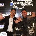 Two men stand and smile with their hands raised in front of a backdrop featuring the ProfitMaster logo and event details. They wear formal attire and seem to be at a promotional event.