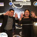 Two people pose playfully in front of a promotional backdrop at the Profitmaster Global Outsourcing event. Both smile and wave, standing on a red carpet.