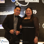 Two people stand together smiling at a formal event with black and white themed decorations in the background. They are both dressed in formal attire.