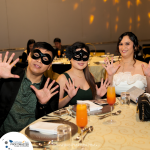 Three people, two wearing black masks and the third not, sit at a table with drinks and pose with raised hands. Fancy event setting with tableware visible. Logo and website text on the tablecloth.