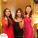 Three women in formal dresses posing and making finger heart gestures in front of a wooden backdrop with gold decorations and balloons. A branded Profittmaster.com.au logo is visible on the bottom left.