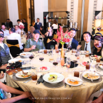 A group of people wearing formal attire and masquerade masks sit around a decorated dinner table at an indoor event. Plates of food and drinks are on the table.