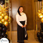 A person stands in front of a decorated background with gold and black balloons, wearing a white top and black skirt. A logo for "Profit Master" is visible in the bottom left corner.