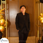 A man in a black suit is standing and smiling in front of a wooden door decorated with black and gold balloons.