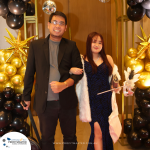 A man and a woman dressed in formal attire stand together, posing for a photo in front of a balloon arch decorated with golden stars and a disco ball. .