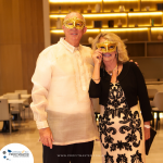 An older couple stands indoors wearing masquerade masks. The man is in a barong tagalog, and the woman is in a black and white dress. The background features shelves with golden lighting.