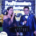 Two individuals, dressed formally, speak into microphones on stage during Profitmaster's 10th Anniversary Gala. A decorated backdrop and music stand are visible.