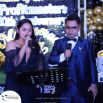 A man and a woman in formal attire speaking into microphones on a stage at the Profitmaster's 10th Anniversary event. They stand in front of a gold-and-black backdrop decorated with balloons.