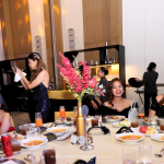 A group of people sitting at a round dinner table with plates of food and drinks in a formal setting. Other guests and decorations are visible in the background.