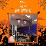 Halloween-themed room with decorations, including a cardboard cutout featuring large eyes and a mustache. "Happy Halloween" text at the top with spider web designs.