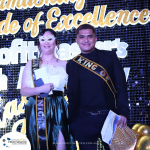 Two people stand on a stage wearing sashes, with the background displaying the words "Pride of Excellence." One wears a mask and holds papers, while the other holds a hat and papers.