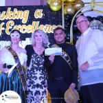 Four people stand on a stage with one holding a "King of the Night" envelope. The background features gold and black balloons, and a sign while the event banner reads "A Masking a Night of Excellence.