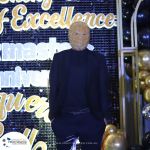 Person wearing a mask with gold-colored beads stands in front of a backdrop with gold and black balloons and "Excellence" sign. A white and blue logo with "PROFITMASTER" is visible in the corner.