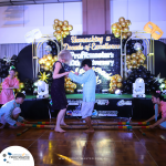 A woman and a child dance on stage while three other children hold a colorful bamboo pole. The background features a balloon arch and a sign reading "Unmasking a Decade of Excellence.