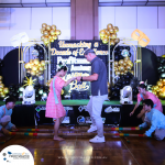 Two people dance on stage surrounded by decorative balloons, while others perform a traditional bamboo dance. A backdrop displays an event theme and sponsor logos.