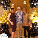 Three people stand together at a formal event with black and gold balloon decorations. Two wear masks, one in a dark dress and the other in casual attire, while the third person is in a traditional outfit.