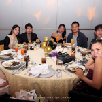 A group of nine people seated around a table, dressed formally, attending an event with food and drinks. Various dishes and glasses seen on the table. The backdrop has a website URL: www.profitmaster.com.au.