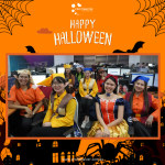 People in Halloween costumes smiling in an office. Decor includes spiders, webs, and bats. Text reads "Happy Halloween" with a company logo above.