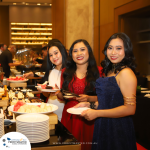 Three women in formal attire stand at a buffet table, serving themselves various food items. The background features warm lighting and stacked plates, indicating a formal event or party setting.