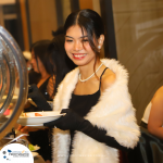 A woman wearing a white shawl and black gloves holds a plate of food at a buffet table. She is smiling and looking down at the plate.