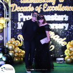 Two people wearing formal attire and masquerade masks smile for a photo. They stand in front of a sparkly backdrop with balloons and text celebrating "Profitmaster's 10th Anniversary.