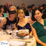 Three women, two wearing masks, sit at a table during a formal event. There are wine glasses and event materials on the table. The background shows seated guests.