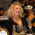 A woman with blonde hair and a black outfit smiles and laughs at a formal event. She is holding a champagne glass, with a masked person in the background.