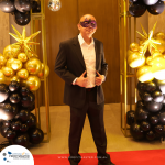 A person in a suit and mask stands on a red carpet bordered by gold and black balloons with star-shaped decorations.